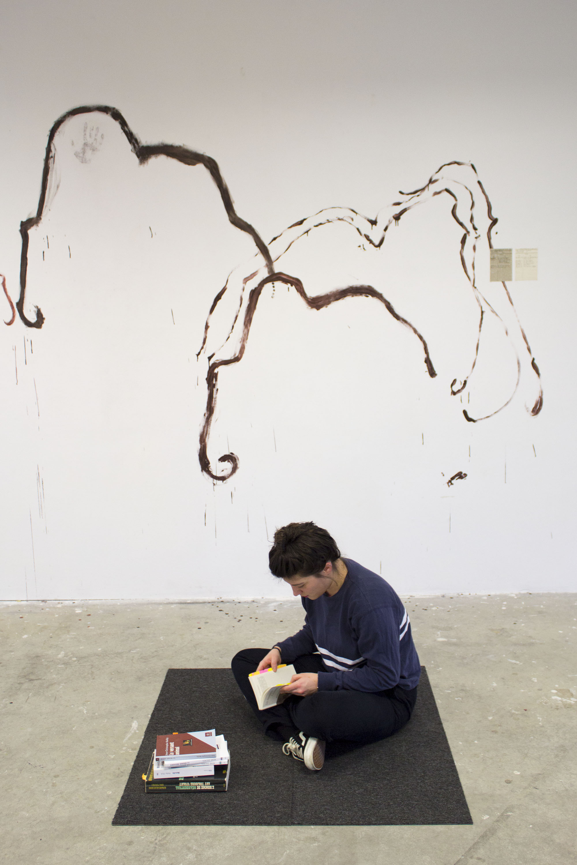Dans une salle avec des dessins de mammouth sur les murs, une jeune fille lit assise sur un carré de moquette. Une pile de livres est posée à côté d'elle. Les livres et la jeune fille ne sont pas les mêmes que précédemment.